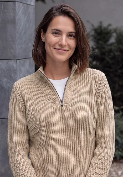 Pull uni à fermeture zip creamy mocha melange Une femme souriante en pull beige et T-shirt blanc devant un décor neutre.