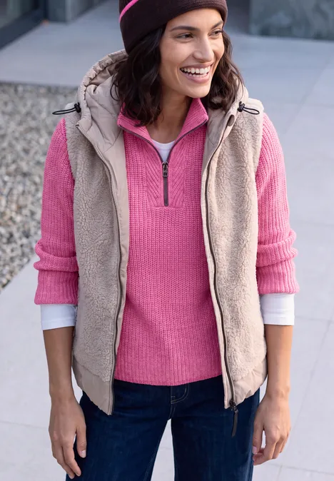 Frau in Jeans mit rosa Pullover, beige Weste und Mütze lächelt im Freien.