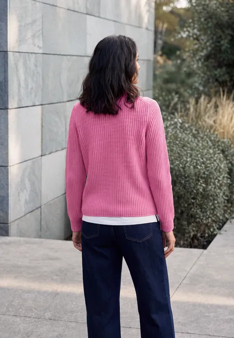Frau in pinkem Pullover und dunkler Jeans steht im Freien, Rücken zur Kamera, vor Mauer und Büschen.