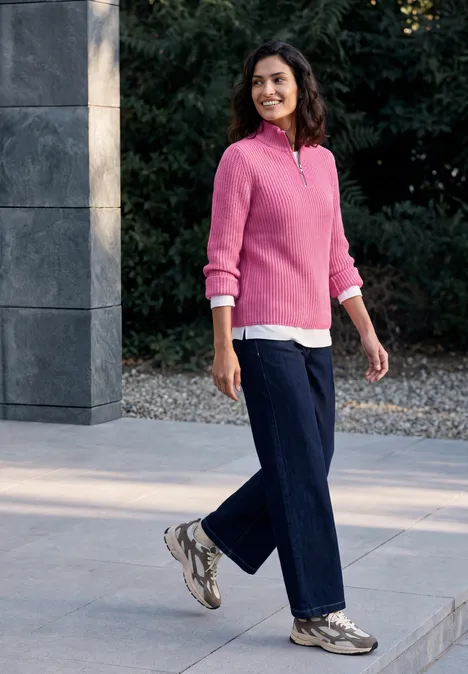Eine Frau in rosa Pullover, Jeans und Sneakers spaziert draußen an moderner Architektur vorbei.