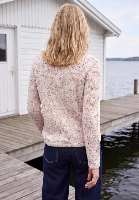 Rückansicht einer Frau in geblümter Bluse und Jeans auf Holzsteg vor weißem Bootshaus am See.