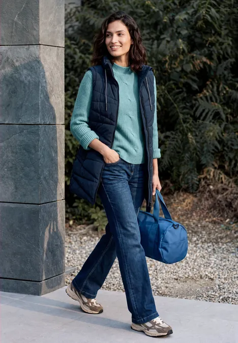 Eine Frau in Weste und Jeans steht draußen an einer Mauer und trägt eine blaue Rundtasche.