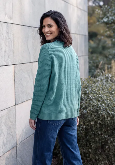 Eine Frau mit lockigen Haaren trägt einen grünen Pullover und blaue Jeans, lehnt lächelnd an einer Steinwand