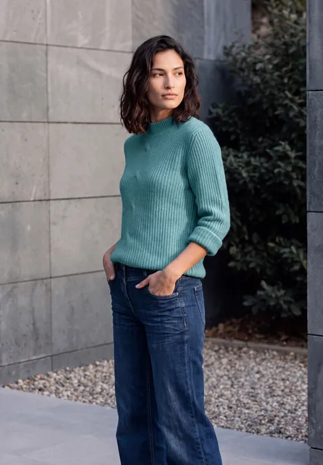 Eine Frau in türkisfarbenem Pullover und Jeans lehnt entspannt mit Händen in den Taschen an einer grauen Wand.