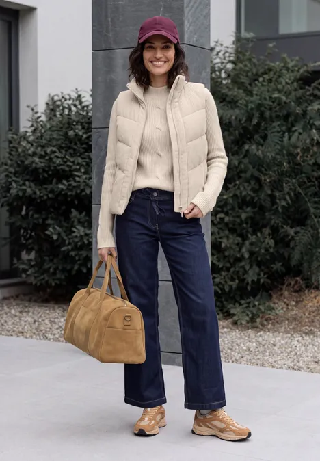 Pull col montant à motif torsadé oat milk beige melange Femme portant une casquette violette, doudoune sans manches beige, pull, jean large, chaussures et sac marron devant un bâtiment.