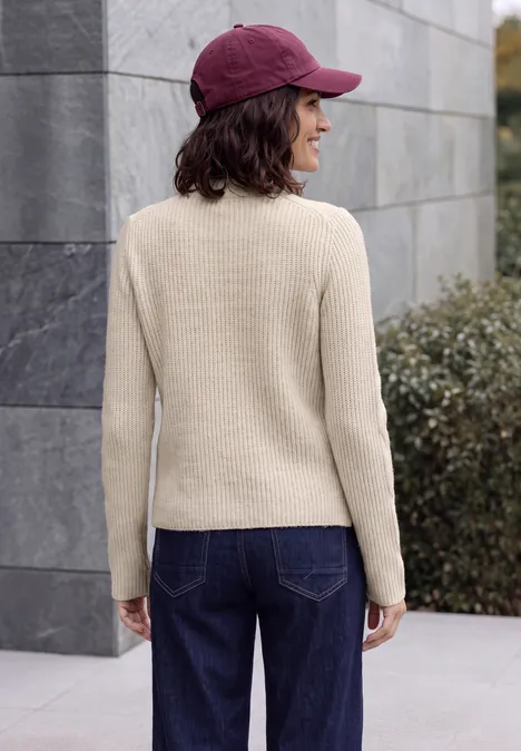 Pull col montant à motif torsadé oat milk beige melange Une femme vue de dos, portant un pull crème, un jean bleu foncé et une casquette bordeaux en extérieur.