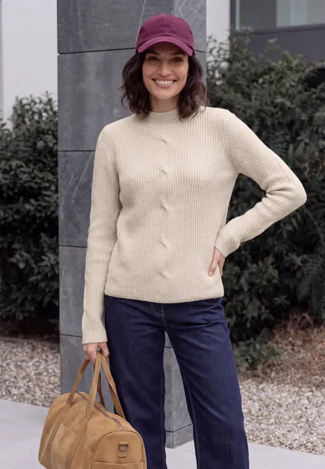 Pull col montant à motif torsadé oat milk beige melange Femme en pull beige, jean bleu et casquette violette, tenant un sac marron, souriante en extérieur.