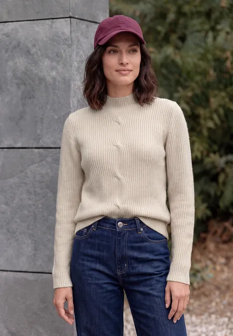 Pull col montant à motif torsadé oat milk beige melange Femme en casquette rouge, pull beige et jean bleu devant un mur en pierre en plein air.