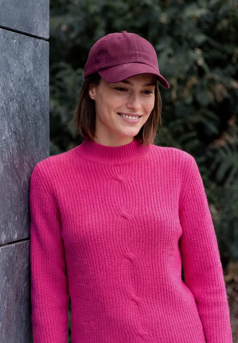 Eine junge Frau lehnt lächelnd an einer Wand, trägt pinken Pullover und Cap.