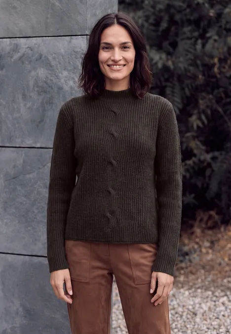 Eine Frau mit dunklem Strickpullover und braunen Hosen steht im Freien und lächelt.