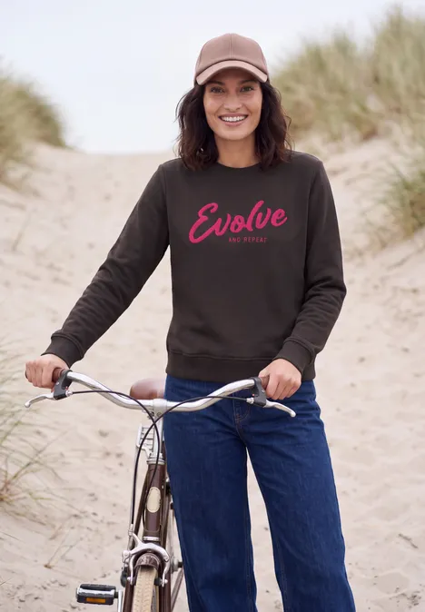 Vrouw met pet en donker 'Evolve'-sweater bij fiets op een zandig pad langs de duinen