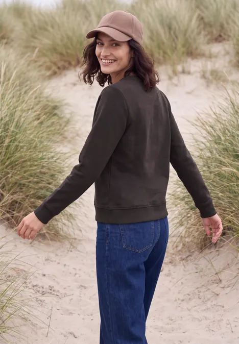 Een glimlachende vrouw in jeans, bruin jack en pet wandelt over de duinen tussen het helmgras.