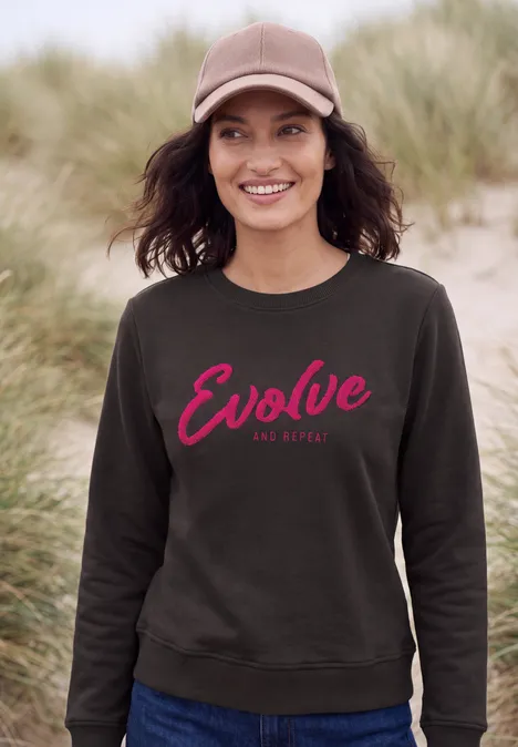 Vrouw in grijze trui met rode 'Evolve'-opdruk, staand bij duinenstrand.