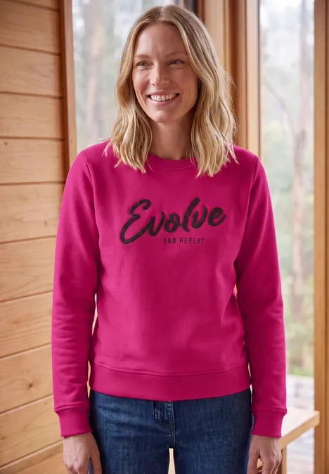 Sweatshirt met ronde hals en tekst pink flash Een lachende vrouw in een roze trui met 'Evolve' tekst, voor een houten raam.