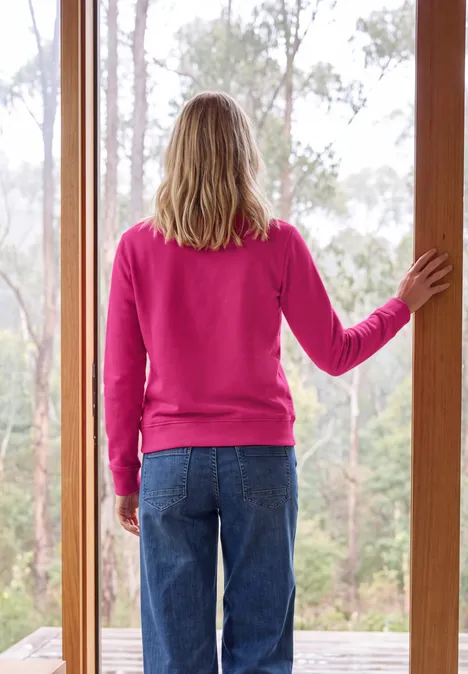 Sweatshirt met ronde hals en tekst pink flash Persoon van achteren in een roze trui en spijkerbroek staat in de deur en kijkt uit op het bos.
