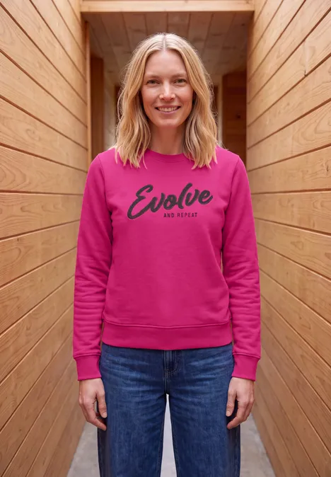 Sweatshirt met ronde hals en tekst pink flash Een glimlachende vrouw in een roze 'Evolve' sweater en spijkerbroek staat in een houten gang.