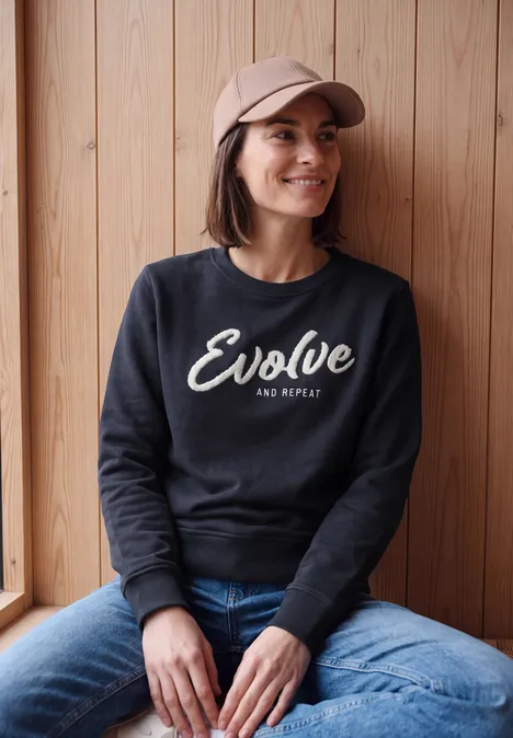 Sweatshirt mit Rundhals und Wording urban dark blue Eine Frau sitzt entspannt vor einer Holzwand, trägt Cap, schwarzen Pullover mit Schriftzug und Jeans.