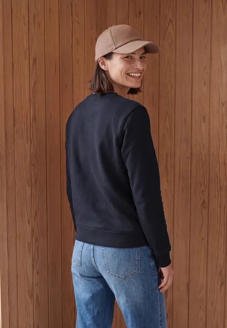 Sweatshirt mit Rundhals und Wording urban dark blue Eine Frau mit Basecap, schwarzem Pullover und Jeans steht vor Holzpaneelen, blickt lächelnd über die Schulter.