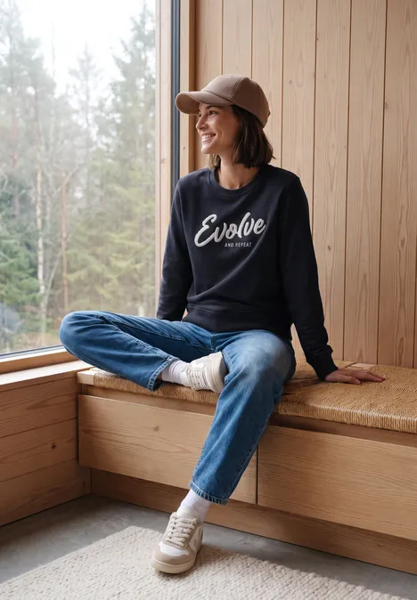 Sweatshirt mit Rundhals und Wording urban dark blue Frau sitzt gemütlich auf Fensterbank in Holzhütte, trägt Jeans, Sneaker und Cap, Pullover mit „Evolve“-Schriftzug.
