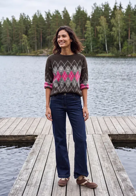 Eine lächelnde Frau steht auf einem Holzsteg am See vor Waldkulisse, sie trägt gemusterten Pullover und Jeans.