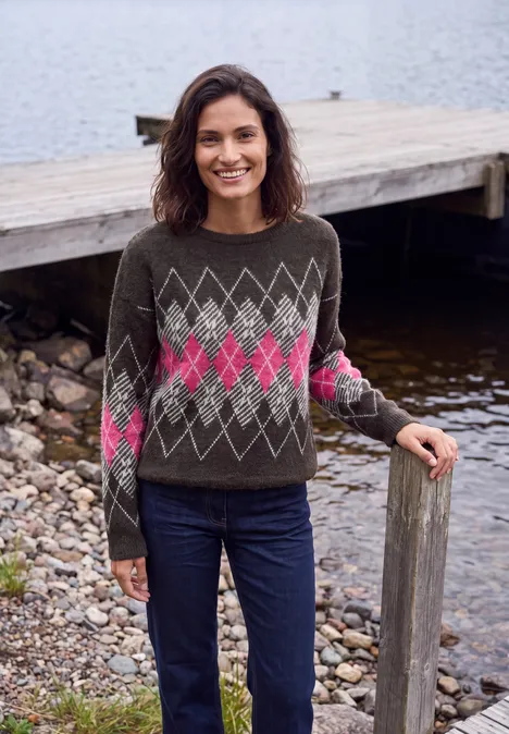 Eine Frau in einem grauen Pullover mit rosa Rautenmuster und Jeans steht lächelnd am Seeufer.