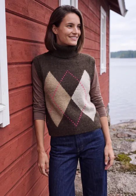 Vest zonder mouwen met kraag en argylepatroon truffle brown Vrouw glimlachend in rolkraagtrui met ruitpatroon, leunend tegen een rood houten huis aan de oever.