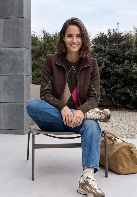 Pull à col rond et motif Argyle truffle brown Une femme souriante assise à l'extérieur, porte jean, pull, gilet et baskets, sac posé à côté.