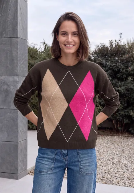 Pull à col rond et motif Argyle truffle brown Une femme souriante posant dehors devant une colonne et des haies, vêtue d’un pull marron à losanges colorés et d’un jean.