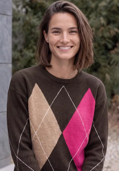 Pull à col rond et motif Argyle truffle brown Femme souriante aux cheveux bruns courts porte un pull marron à motif losanges beiges et roses, debout en extérieur.