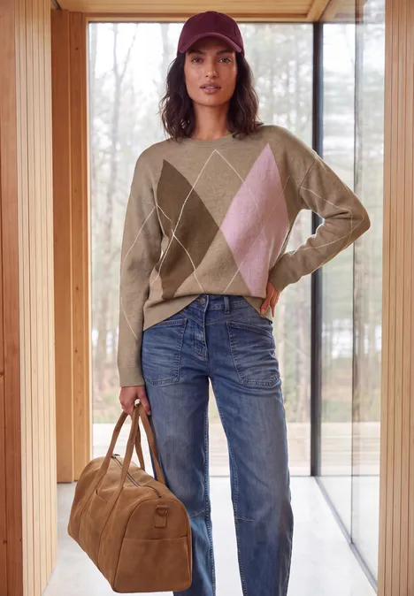 Pullover mit Rundhals und Argyle-Muster creamy mocha melange Frau mit roter Kappe, beigem Rautenpullover, blauen Jeans und brauner Tasche in hellem Raum vor Fenster.
