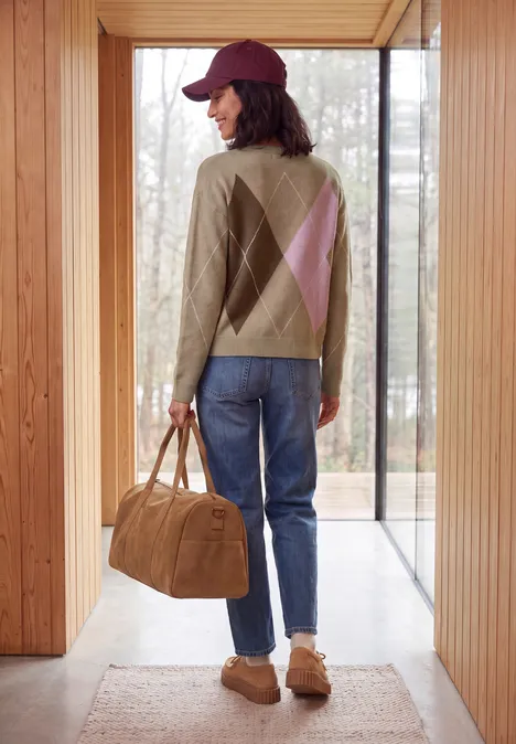 Pullover mit Rundhals und Argyle-Muster creamy mocha melange Eine Person in Mütze, gemustertem Pullover, Jeans und Hausschuhen trägt eine Reisetasche in einem modernen Holzhaus.