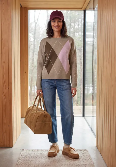 Pullover mit Rundhals und Argyle-Muster creamy mocha melange Frau steht in einer Holzrahmen-Tür, trägt argyle Pullover, Jeans, Cap und hält eine große Umhängetasche.