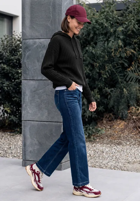 Eine Frau in roter Mütze, schwarzem Hoodie und Jeans spaziert lächelnd an einer Mauer entlang.