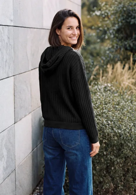 Eine Frau in schwarzem Kapuzenpullover und Jeans lehnt im Freien an einer Wand und lächelt.