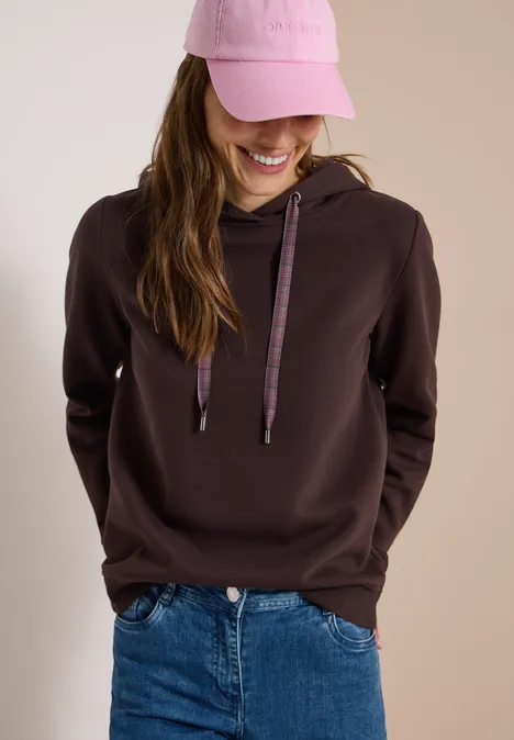 Junge Frau lächelt, trägt braunen Kapuzenpullover, blaue Jeans und rosa Baseballcap.