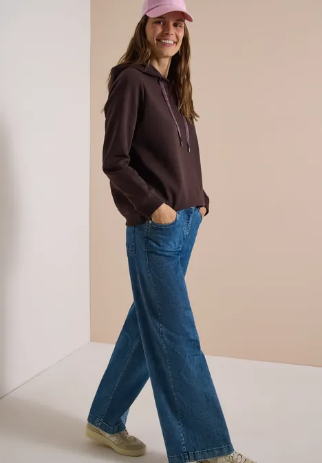 Frau mit rosa Cap, braunem Hoodie und weiten Jeans lehnt locker an Wand und lächelt.