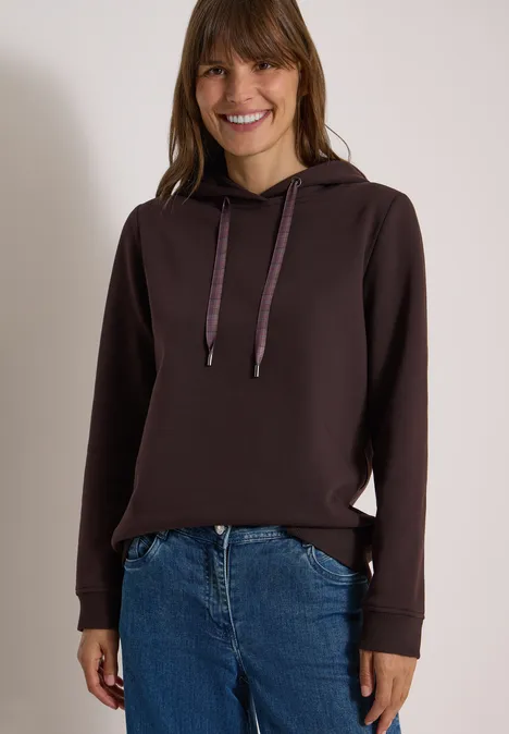 Eine lächelnde Frau trägt einen dunkelbraunen Kapuzenpullover und Jeans vor hellem Hintergrund.