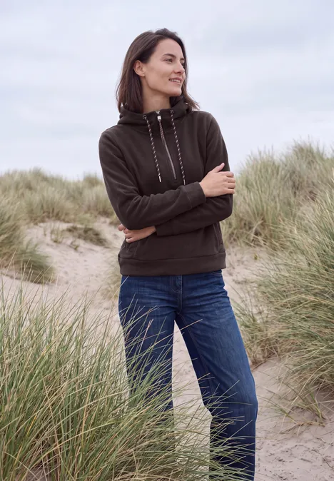 Een vrouw in een hoodie en spijkerbroek met gekruiste armen staat op grasduinen bij het strand.