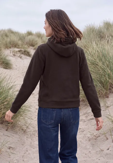 Iemand van achteren in een bruine hoodie en jeans, lopend door duingras op het strand.