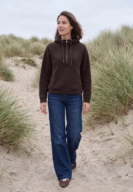 Vrouw in zwarte hoodie en blauwe jeans loopt over met gras begroeide zandduinen op het strand.