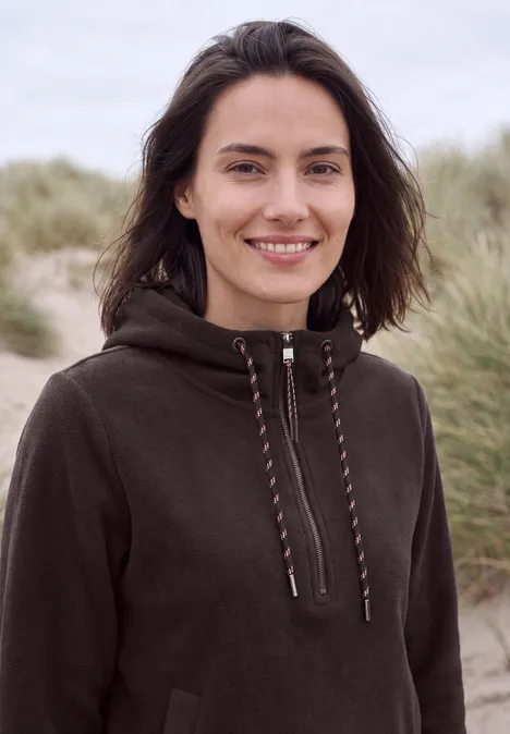 Glimlachende vrouw in een donkere capuchonjas, staand op een strand voor duinen.