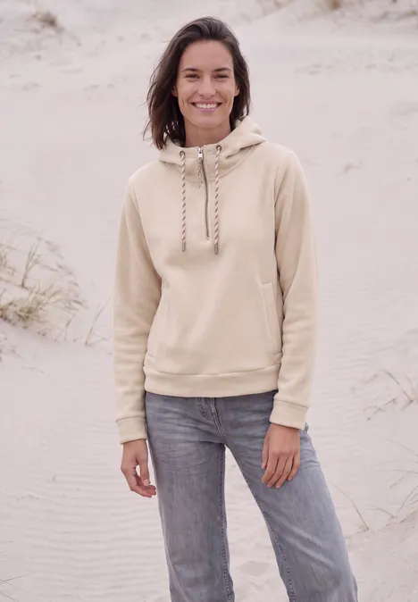 Eine lächelnde Frau in beigem Hoodie und Jeans steht am Strand im Sand.