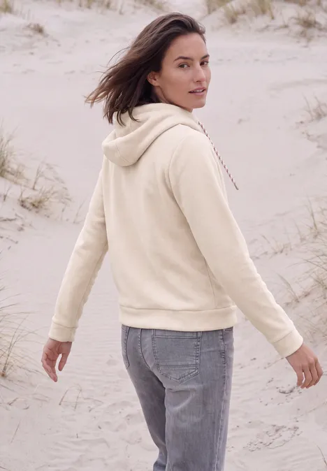 Eine Frau in beiger Kapuzenjacke und Jeans steht auf Sanddünen und blickt über die Schulter.
