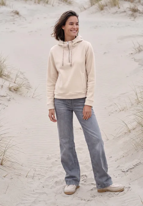 Eine Frau in beigem Hoodie und weiten Jeans steht lächelnd am Strand mit Sanddünen.