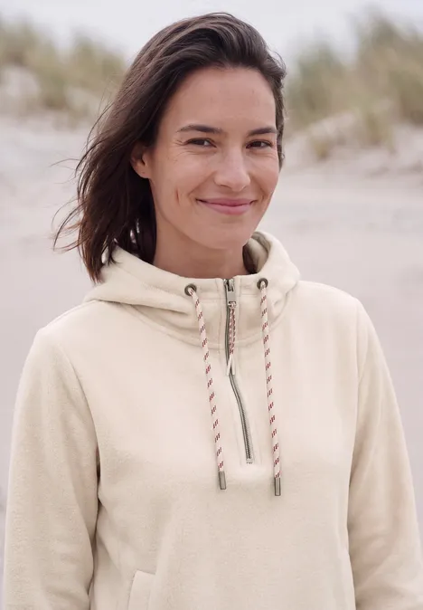 Eine lächelnde Frau mit hellbeigem Kapuzenpullover am Strand im Wind.