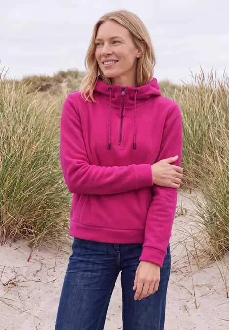 Frau in pinker Kapuzenfleecejacke und Jeans steht lächelnd am Strand vor Dünen mit Strandgras.