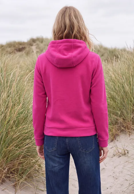 Frau im pinken Hoodie und Jeans steht mit dem Rücken zur Kamera in einer Dünenlandschaft.
