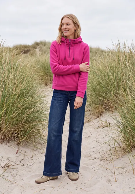 Frau in pinkem Hoodie und Jeans steht lächelnd am grasigen Strand unter bewölktem Himmel.