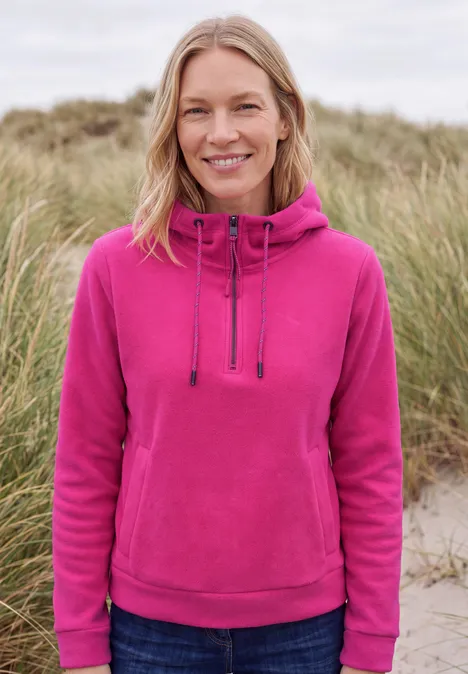 Eine lächelnde Frau mit blonden Haaren trägt einen pinken Kapuzenpullover und Jeans am Strand.