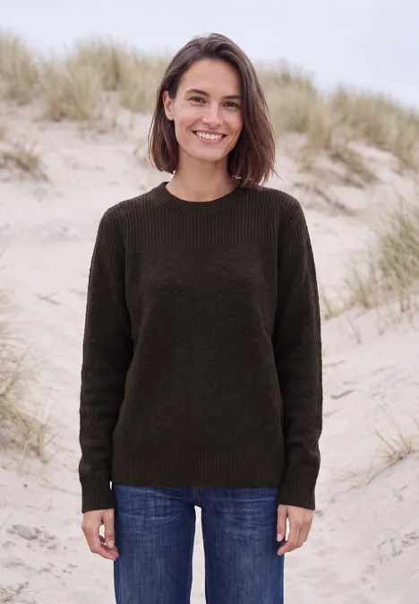 Trui met bouclé-mix en ronde hals truffle brown melange Een vrouw in een donkere trui en spijkerbroek lacht voor grasrijke duinen.